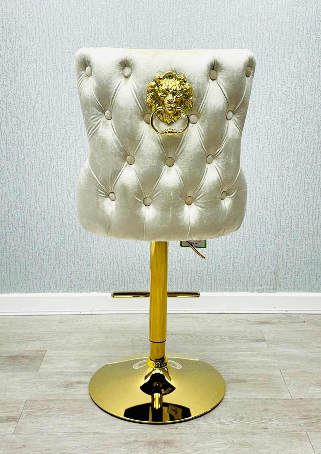 A cream colored bar stool with gold accents, featuring a tufted backrest and a lion knocker on the back.