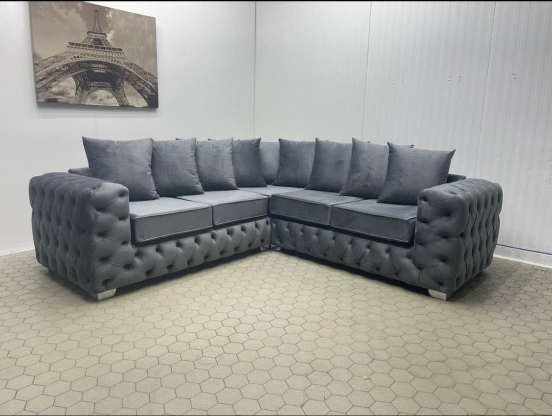 A dark grey, corner sofa with a scatter back, made of plush velvet, featuring tufted upholstery and armrests.