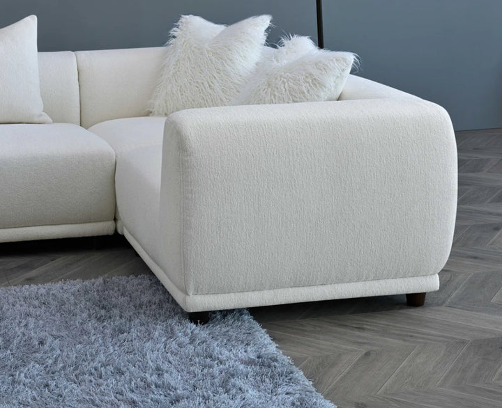 A white modular corner sofa with multiple cushions in a living room setting, complemented by a wooden frame and a plush grey rug.