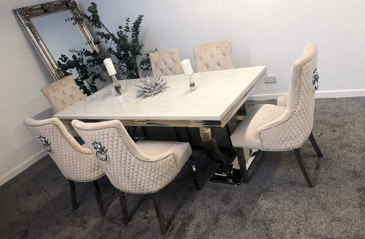 A white marble dining table with a stainless steel base accompanied by beige upholstered dining chairs with tufted backrests and lion knocker accents.