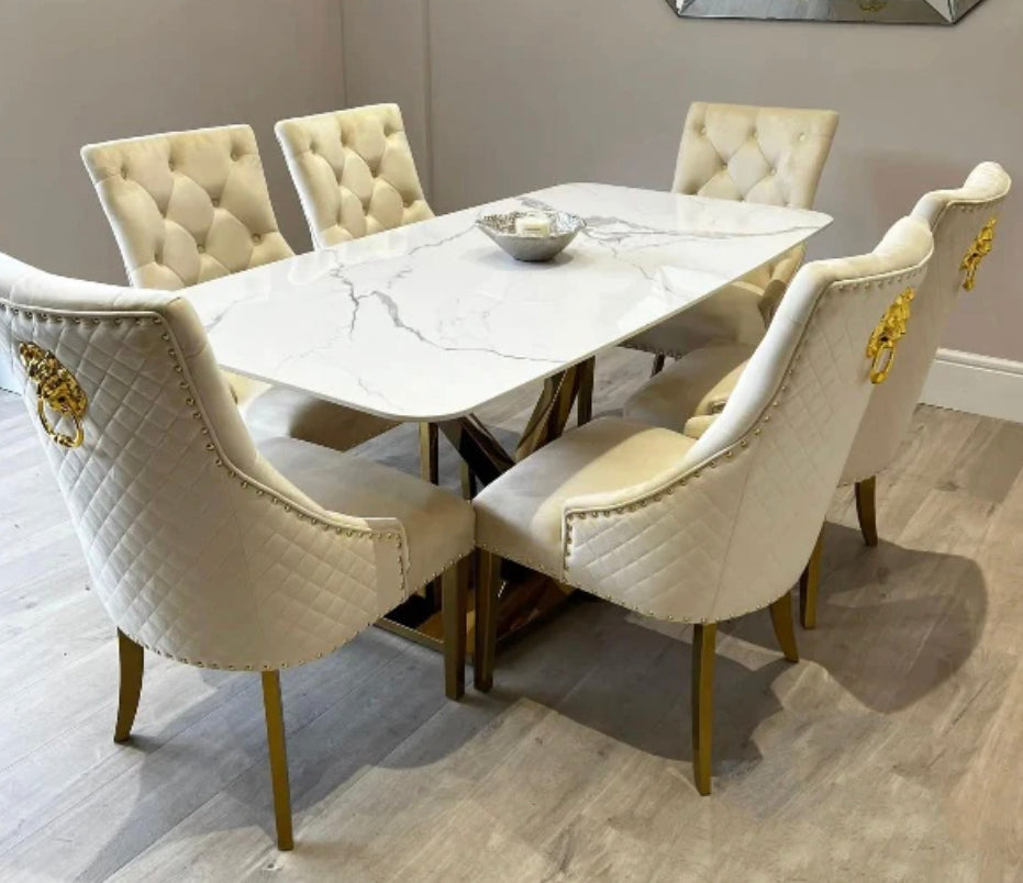 A white marble dining table with a gold-colored metal leg base, accompanied by six cream upholstered dining chairs with gold lion knocker designs.