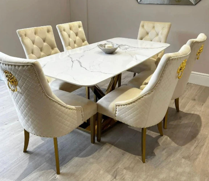 A white marble dining table with a gold-colored metal leg base, accompanied by six cream upholstered dining chairs with gold lion knocker designs.