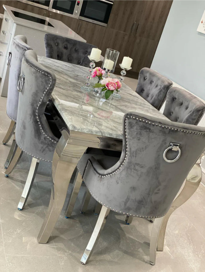 A grey marble dining table with stainless steel legs, surrounded by dark grey plush velvet chairs with chrome ring knockers.