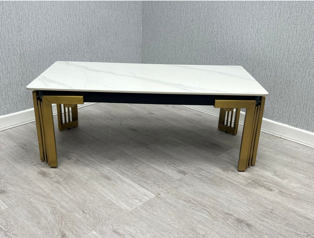 A white ceramic coffee table with a gold frame and gold accents on a grey floor, with a marble-like appearance on the tabletop.