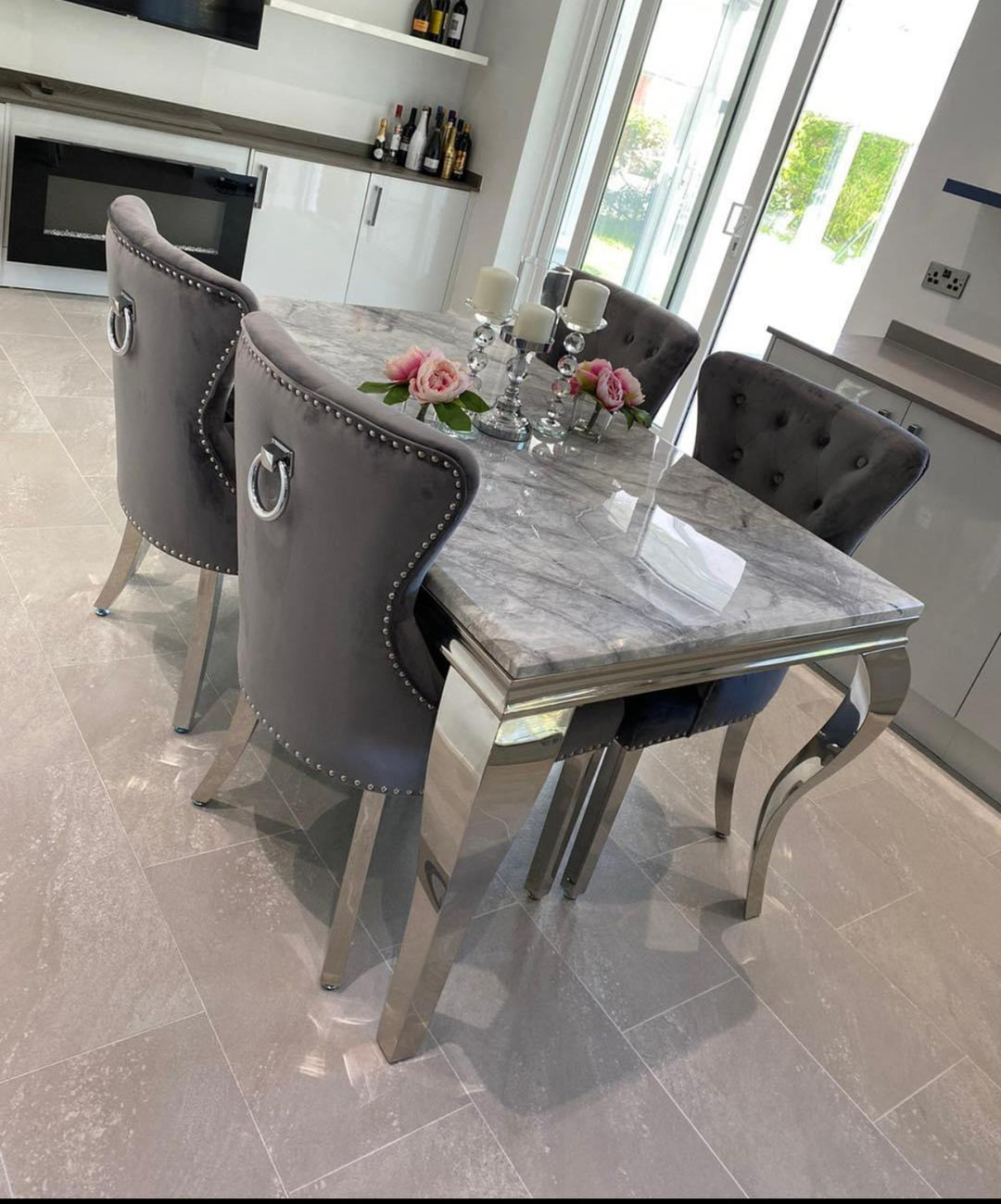 A light grey marble dining table with silver legs, accompanied by dark grey chairs with chrome ring knocker handles, set in a well-lit room.