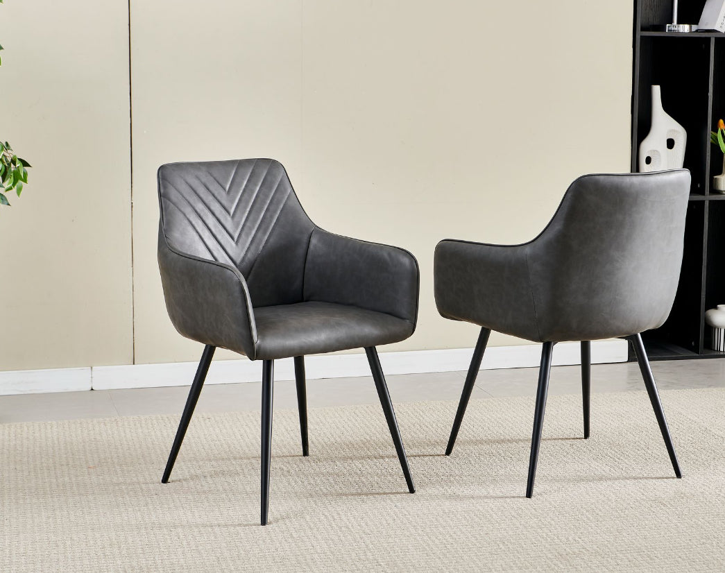 Pair of modern dark grey faux leather dining chairs with chevron-stitched backrest, black tapered legs, and curved armrests on a beige rug