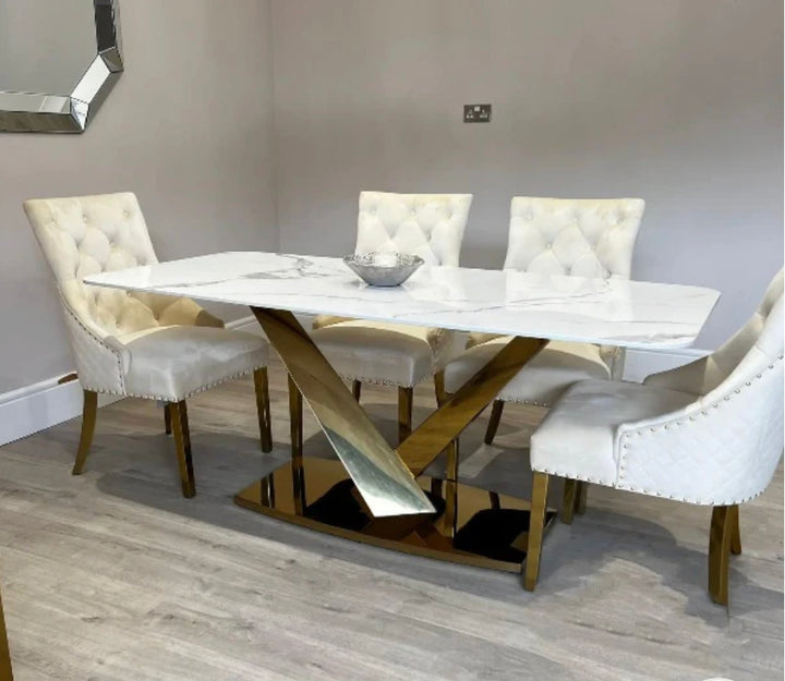 A white marble dining table with a gold-colored metal leg base, accompanied by six cream upholstered dining chairs with gold lion knocker designs.