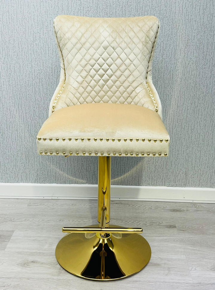 A cream colored bar stool with gold accents, featuring a tufted backrest and a lion knocker on the back.