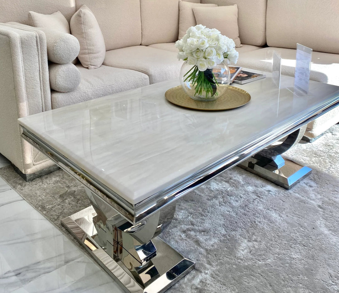 A chic cream-colored marble coffee table with a stainless steel double circular base, positioned in a room with a white sofa and a round rug.