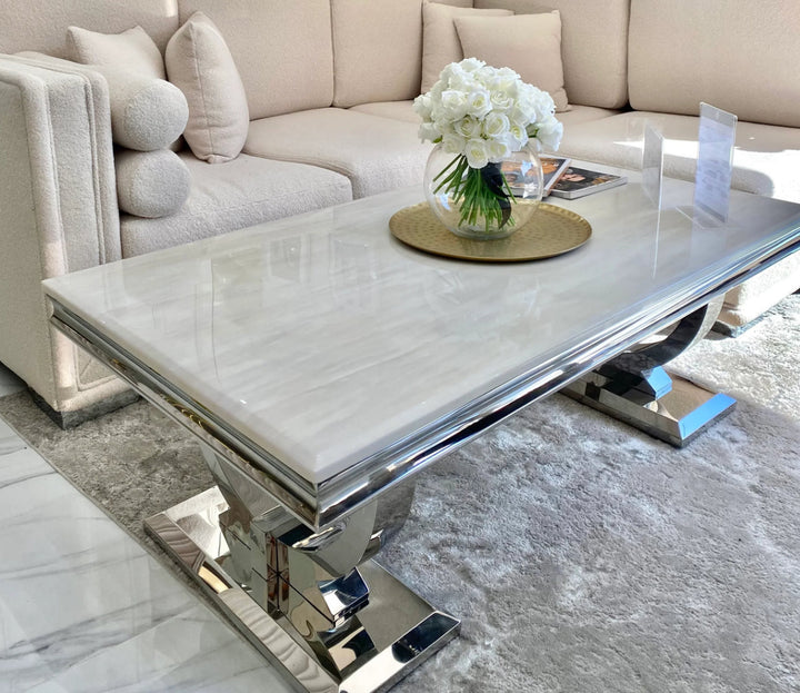 A chic cream-colored marble coffee table with a stainless steel double circular base, positioned in a room with a white sofa and a round rug.