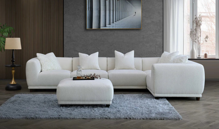 A white modular corner sofa with multiple cushions in a living room setting, complemented by a wooden frame and a plush grey rug.