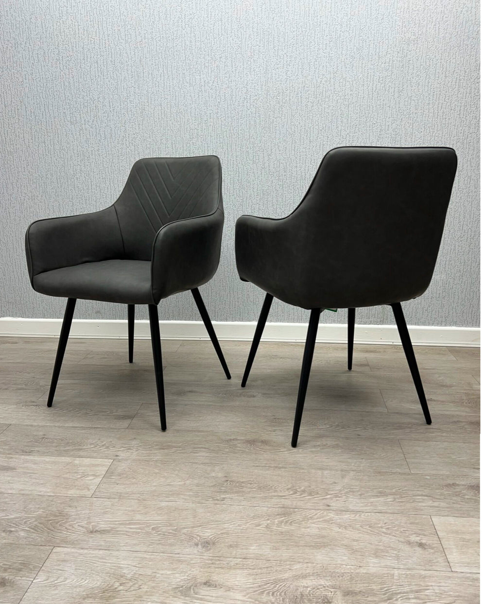 Two gray faux leather dining chairs with armrests and metal legs on a wooden floor.