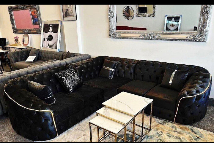 A large black corner sofa with gold accents, arranged in a showroom setting with decorative cushions and a coffee table in the foreground.
