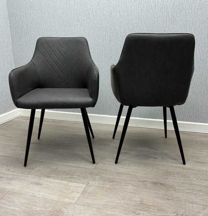 Two gray faux leather dining chairs with armrests and metal legs on a wooden floor.