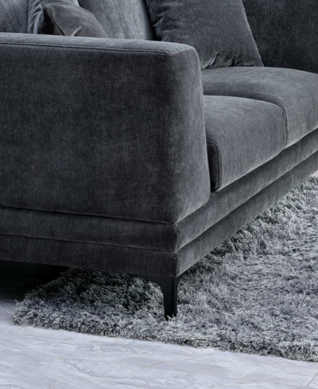 Close-up of dark grey velvet sofa armrest with plush cushions and black metal legs on a soft grey shaggy rug