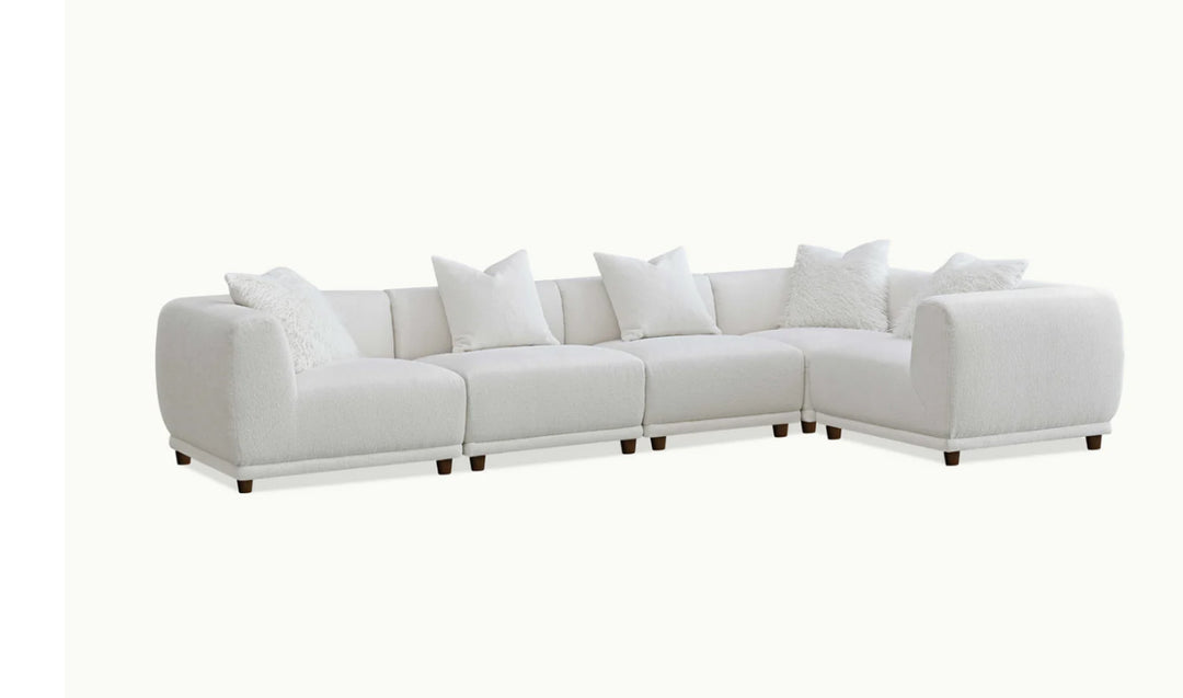 A white modular corner sofa with multiple cushions in a living room setting, complemented by a wooden frame and a plush grey rug.