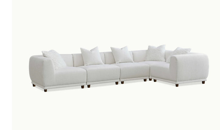 A white modular corner sofa with multiple cushions in a living room setting, complemented by a wooden frame and a plush grey rug.