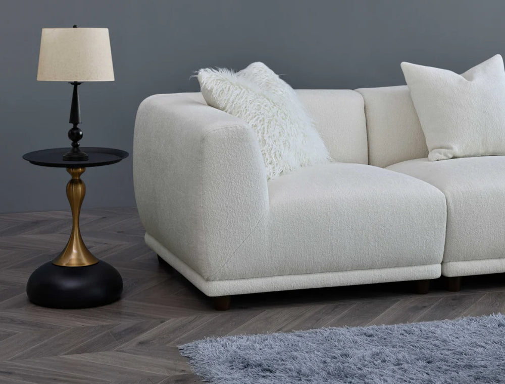 A white modular corner sofa with multiple cushions in a living room setting, complemented by a wooden frame and a plush grey rug.
