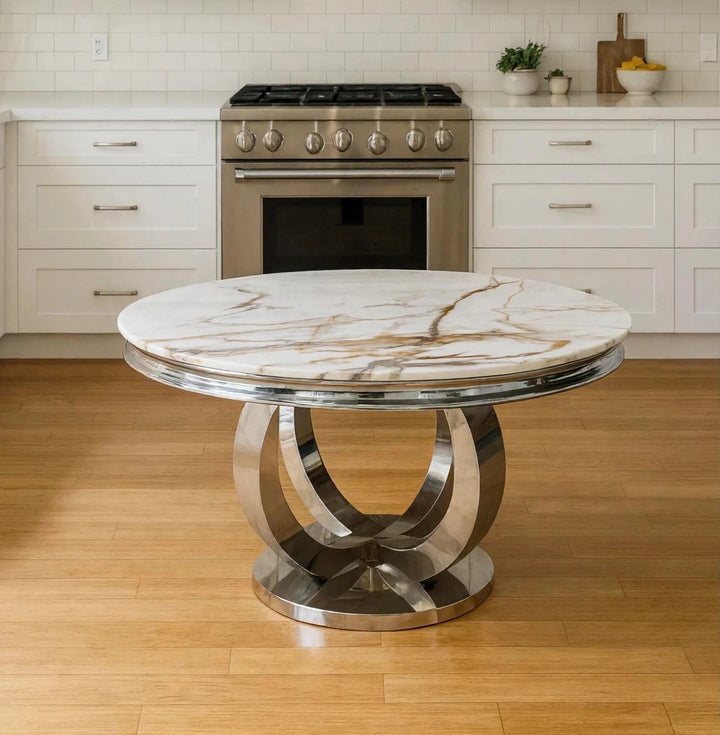 A round dining table with a white and gold veined marble top and a polished chrome curved pedestal base, set in a modern kitchen with white cabinets and light wood floors.