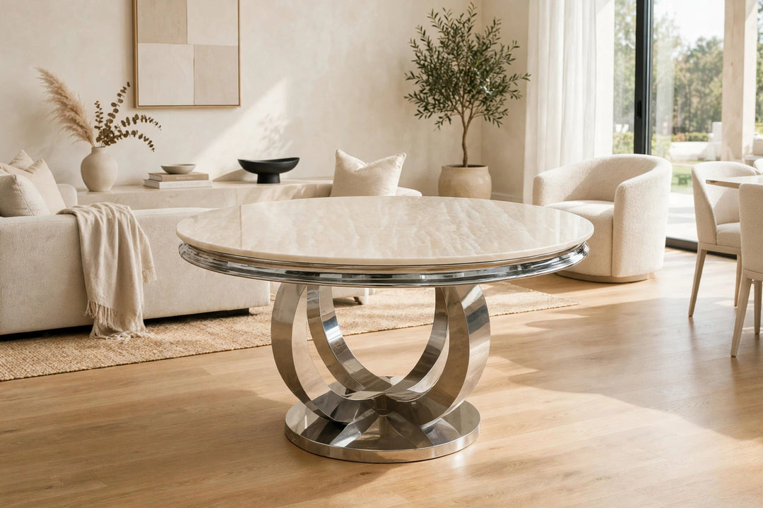 A round dining table with a polished chrome pedestal base and Cream  marble top, set in a bright, modern living room with neutral-toned furniture.