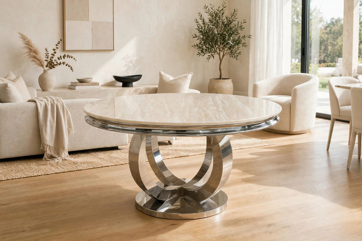 A round dining table with a polished chrome pedestal base and Cream  marble top, set in a bright, modern living room with neutral-toned furniture.