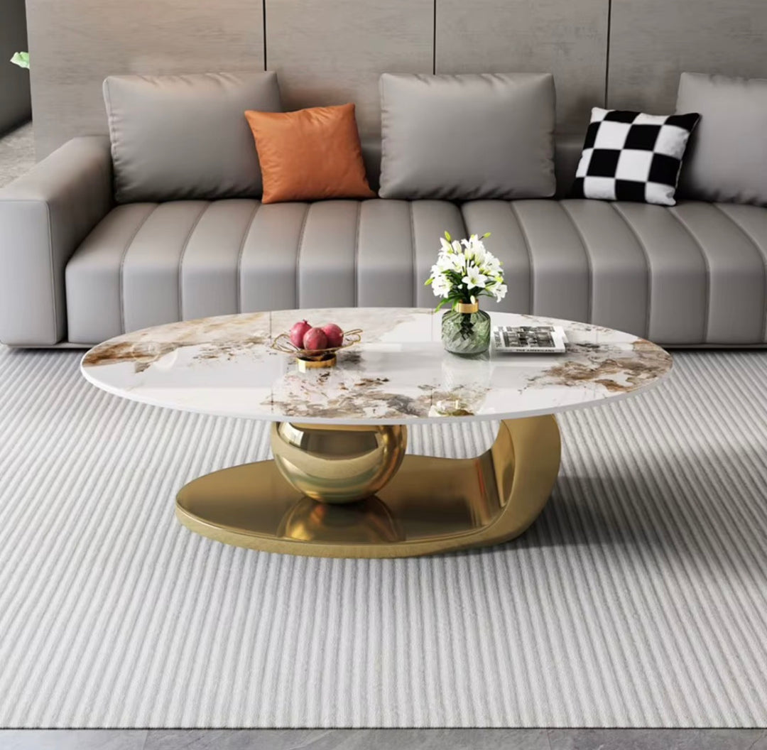 An oval coffee table with a white marble-patterned top and a polished gold base, styled in a modern living room setting in front of a gray sofa.