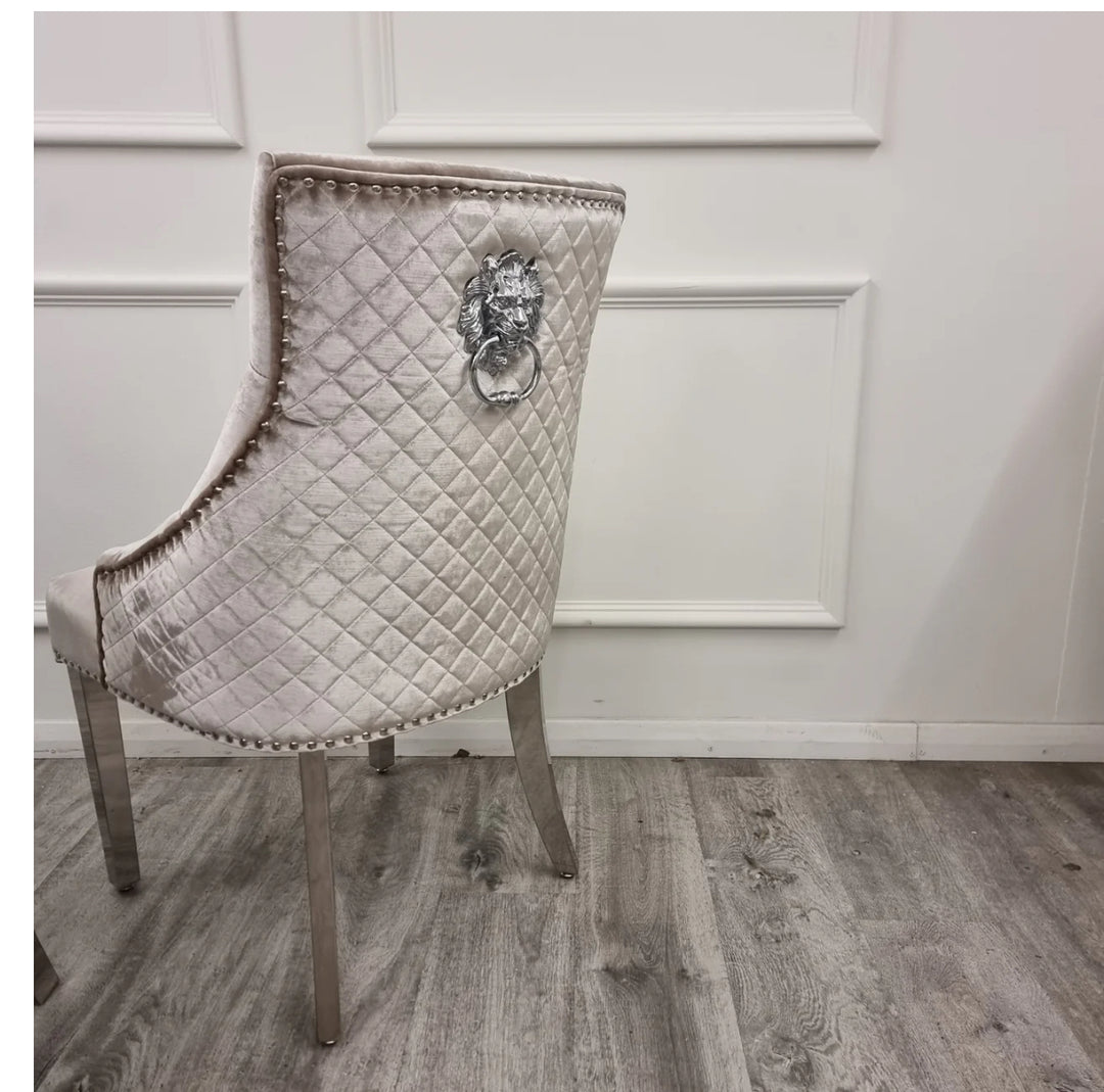 A beige upholstered dining chair with a quilted stitching back design, featuring a polished steel chrome detailed lion knocker and studding detailing along the border. The chair is displayed against a white panel background.